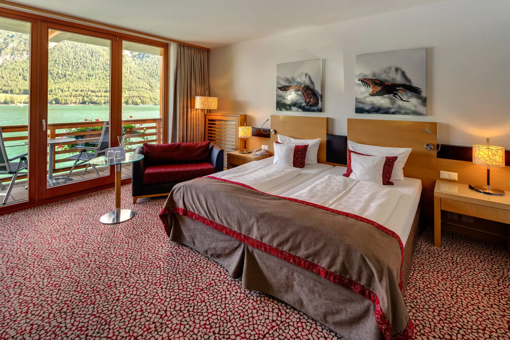 Interior view of a hotel room with bed, chair and table. A balcony with a view of the lake can be seen in the background.