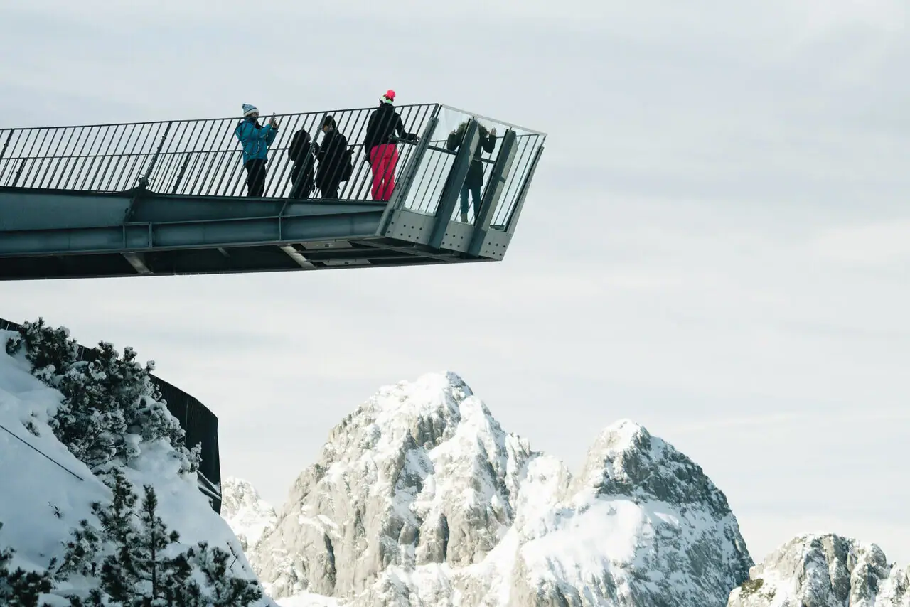 AlpspiX People stand on a bridge over a snow-covered mountain.