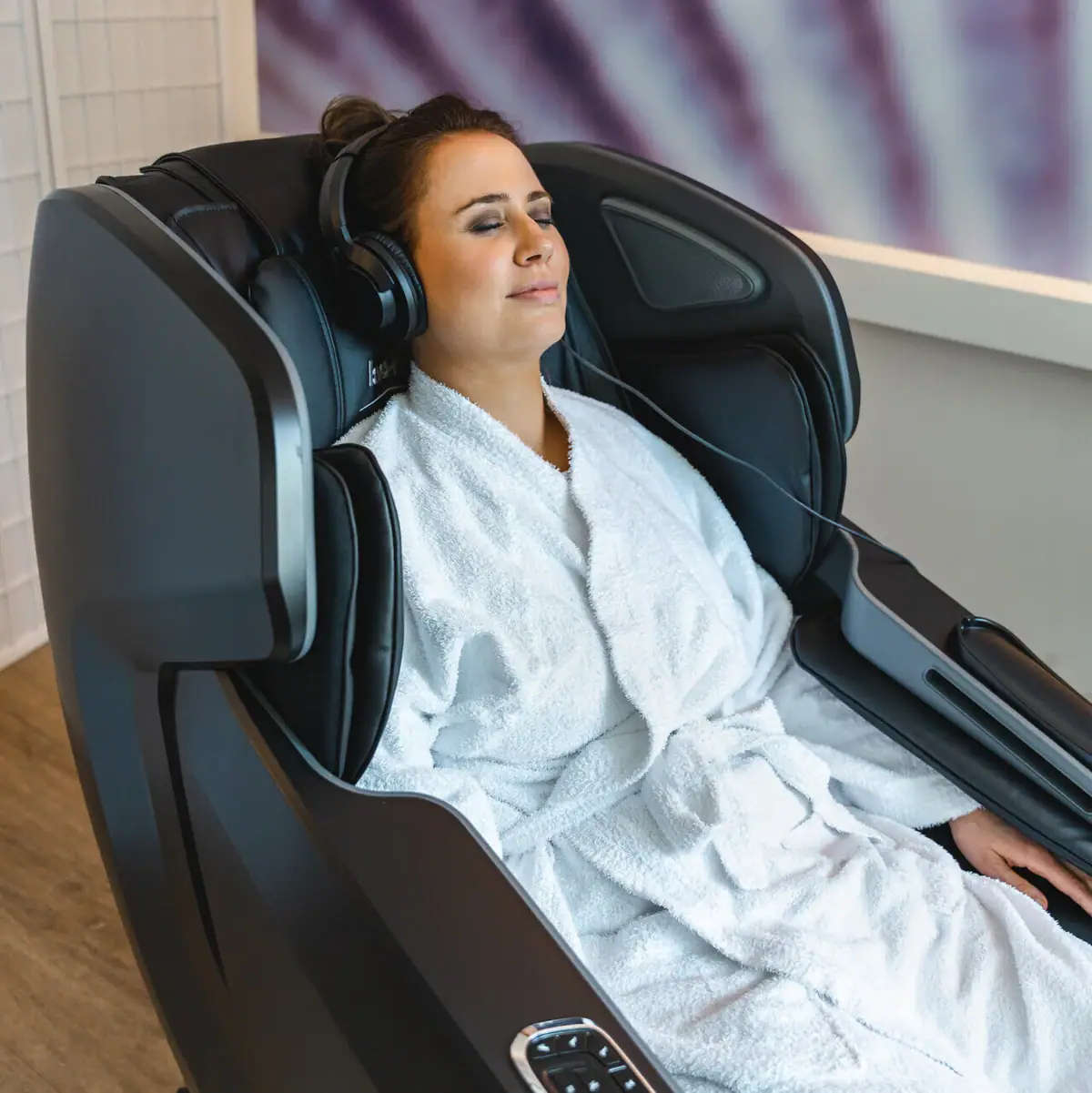 A woman in a bathrobe and headphones sits in a massage chair.