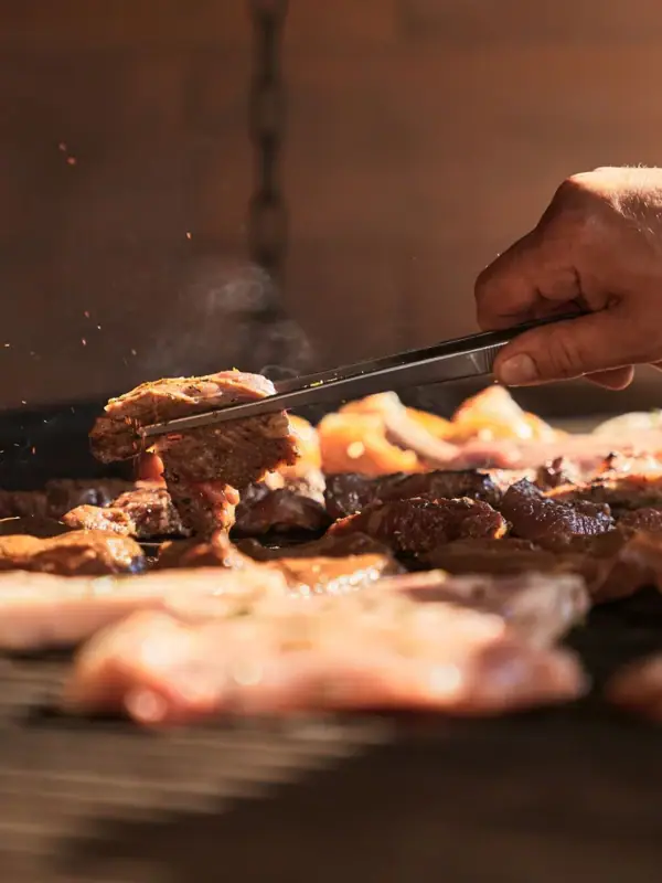 A person holds tongs over meat on a grill.