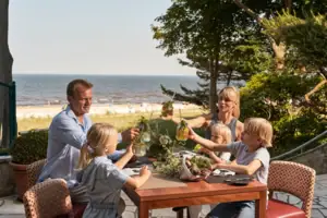 Eine Familie mit 2 Kindern sitzt an einem Tisch mit Getränken und Blick auf das Meer.