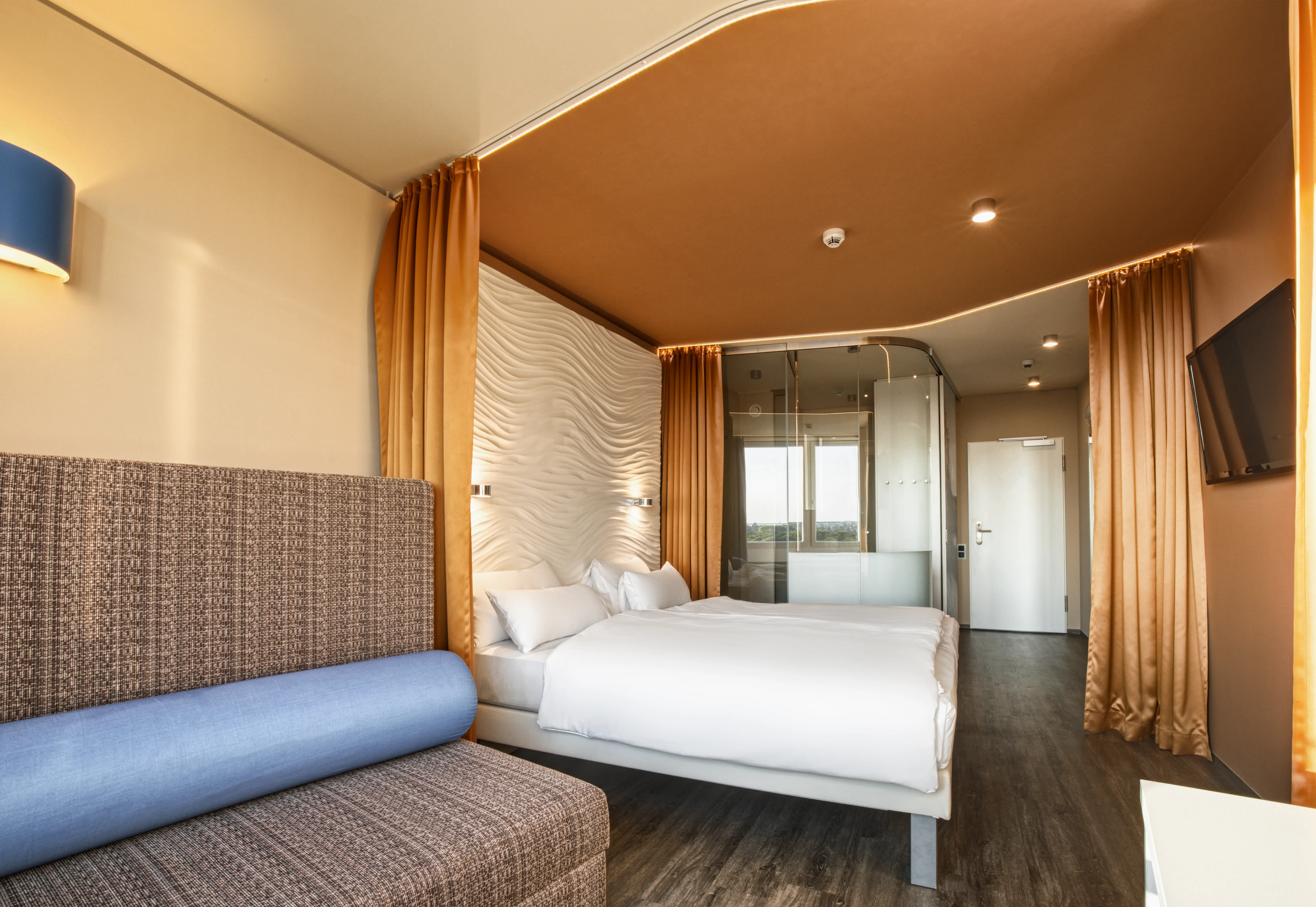 Stylish hotel room featuring a comfortable double bed with a wave-patterned headboard, warm mustard-colored curtains, a seating area with a blue cushion, and an open-plan bathroom with glass partitions, creating a modern and inviting ambiance.