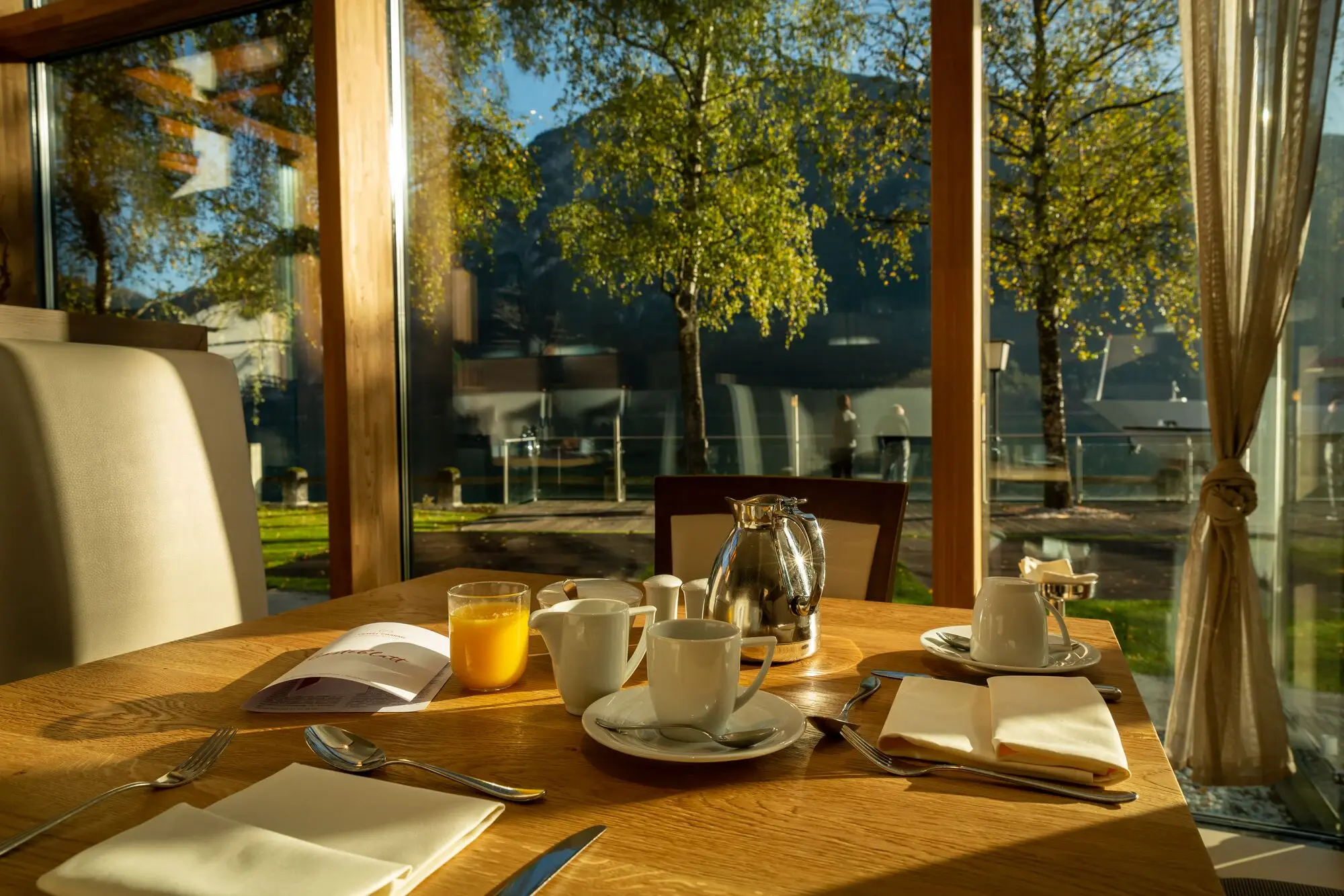A table with a glass teapot, teacups and saucers at a table next to a window with a view of trees.