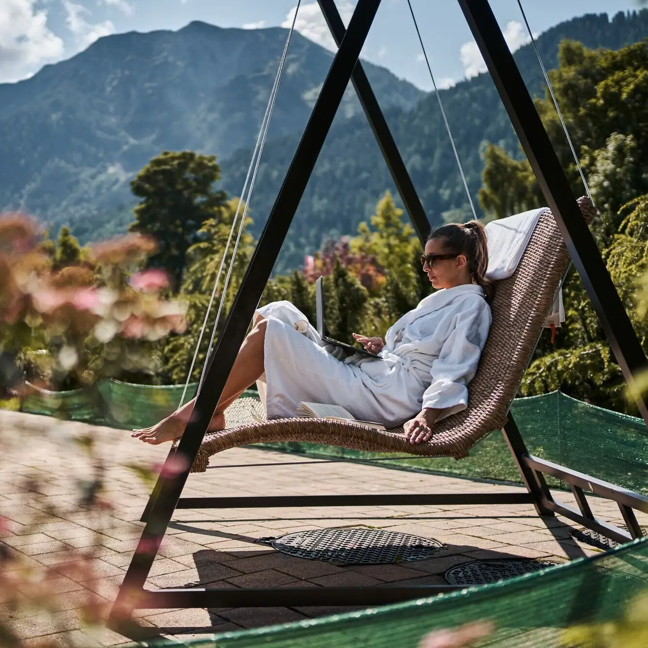A woman in a white bathrobe sits in a hammock with a laptop.