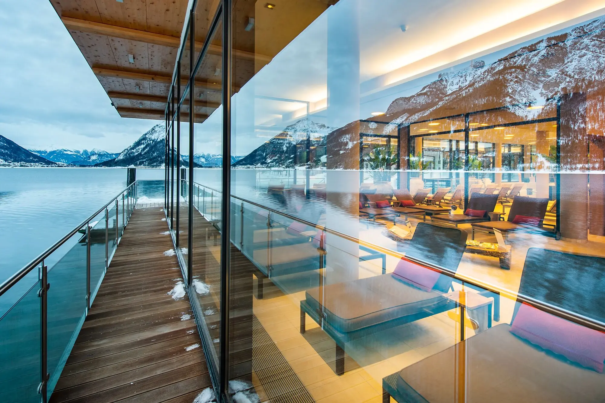 Glass wall of a building with terrace and chairs, view of the Achensee and mountains.