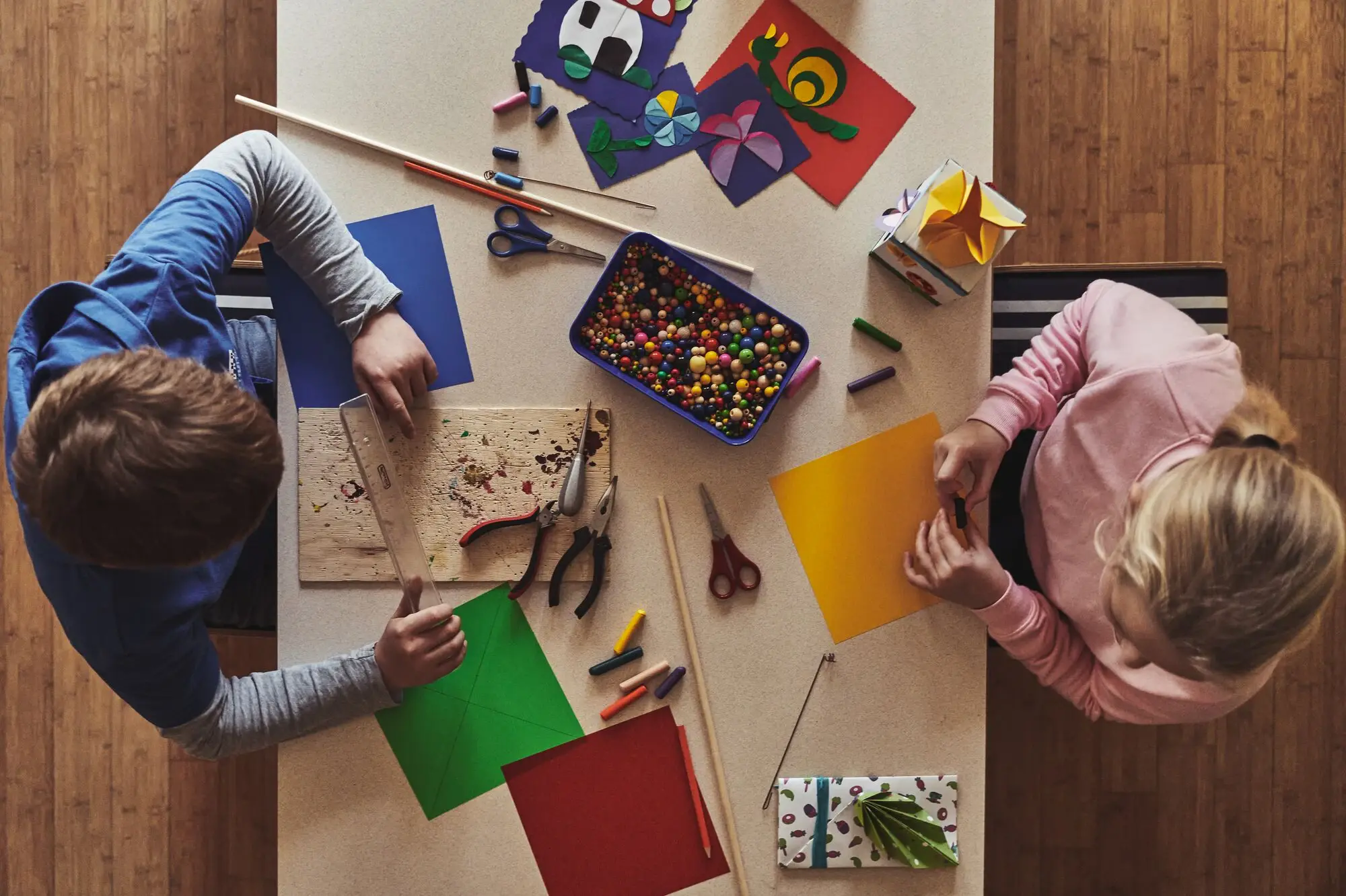 Two children sit at a table and work with paper and craft materials in an interior space.