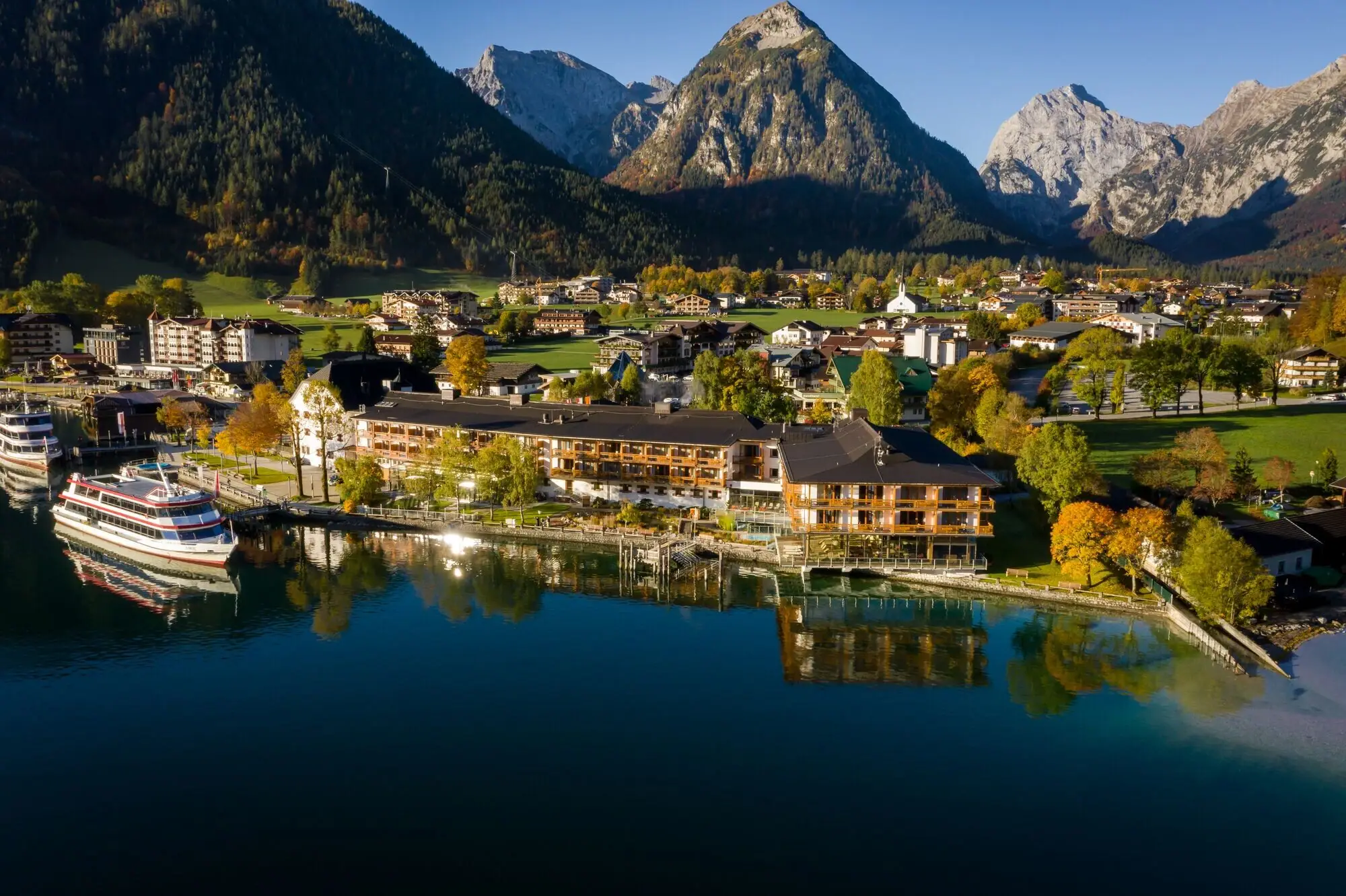 aja Achensee Hotel in front of a mountain panorama A town on the water with trees and a boat in the foreground.
