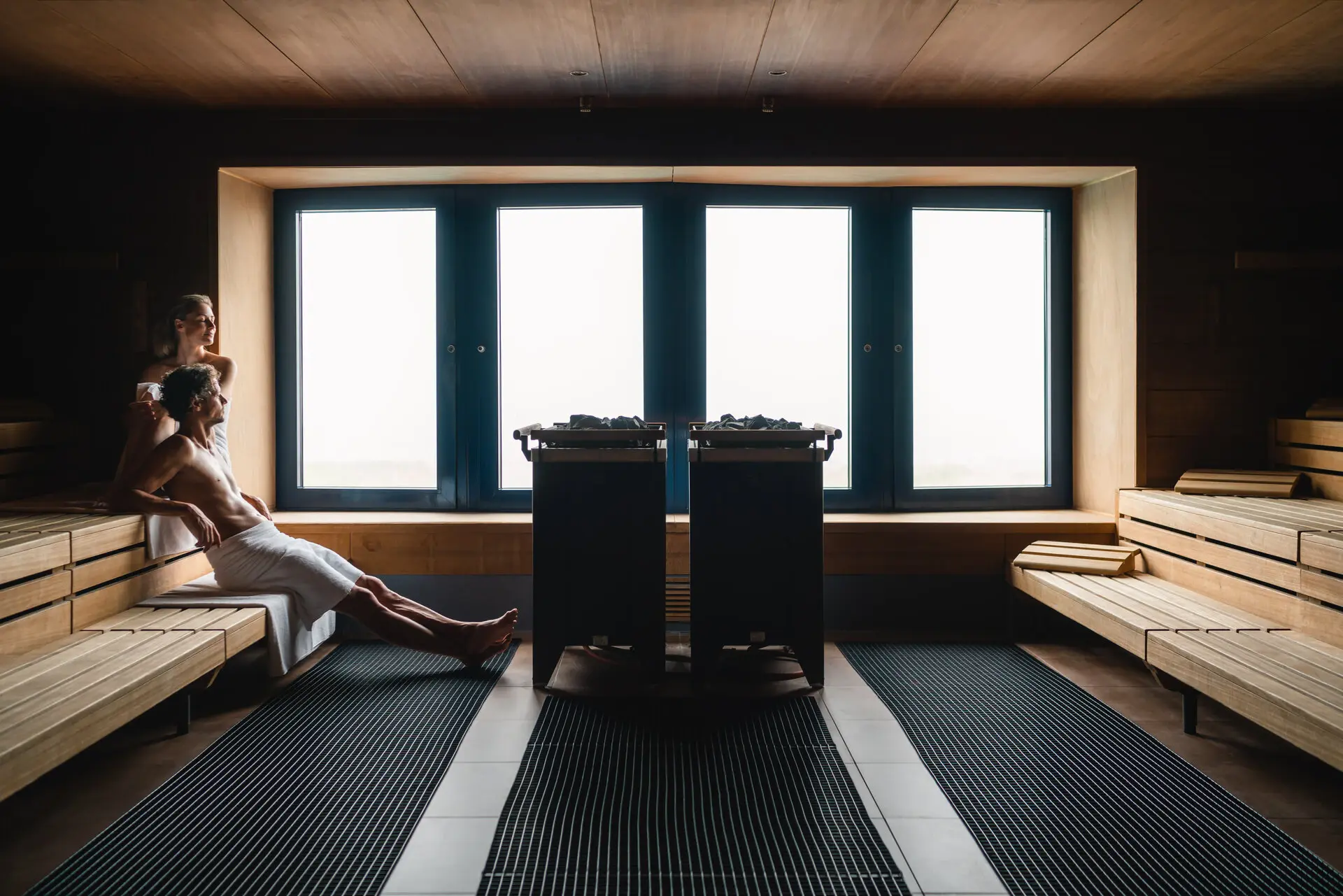 A person sits relaxed on a wooden bench in a sauna in front of a window in the background