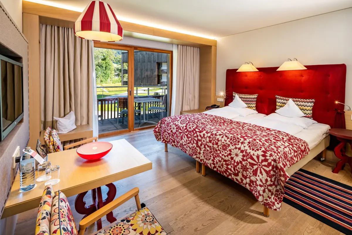Bright and colourful Alpine-style hotel room with double bed, red headboard, patterned bedspread, seating area and access to the balcony with a view of the greenery.