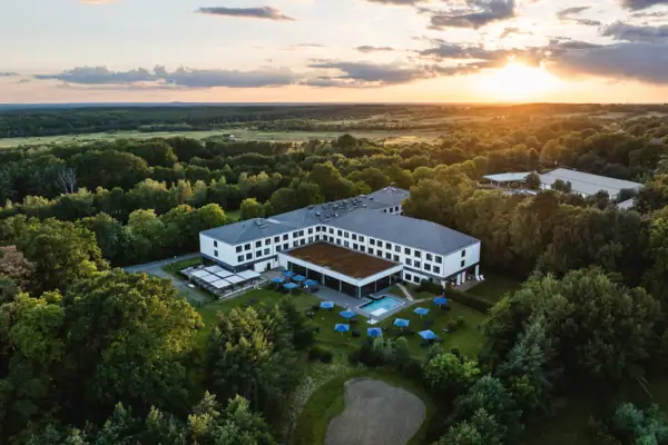 An aerial view of the aja Bad Saarow with outdoor pool in green nature, surrounded by trees at sunset. 