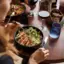 A woman brings her fork with her left hand to a plate of chicken salad on a wooden table.