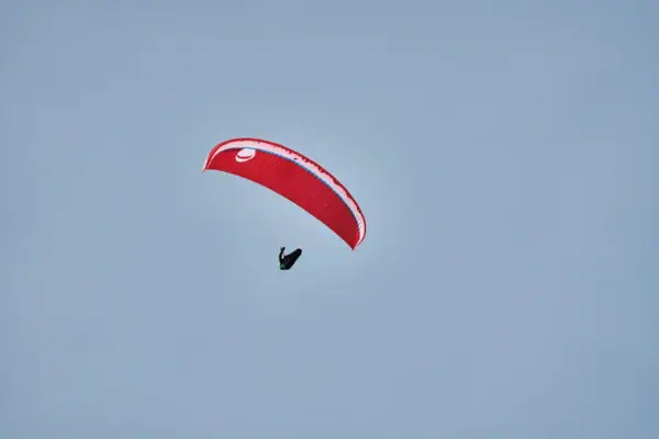 A person paragliding with a red paraglider hovers in front of the blue sky.