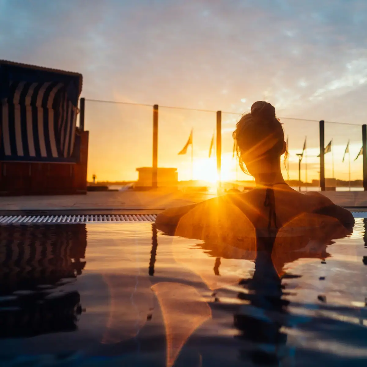 A woman is leaning on the edge of an outdoor pool at sunset, gazing into the distance. The pool is bordered by a glass fence, with the sea behind it. In front of it is a blue and white striped beach chair.