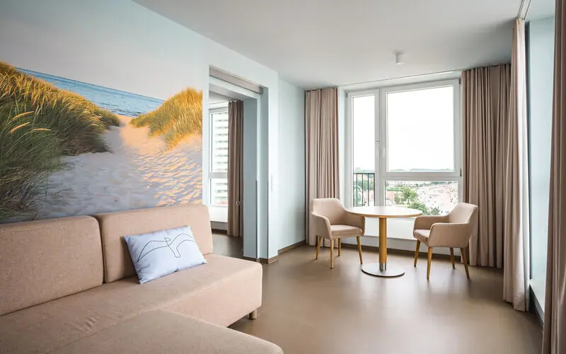 A spacious suite with a sandy dune mural, a beige sofa, a small dining area with two chairs, and large windows providing scenic views.