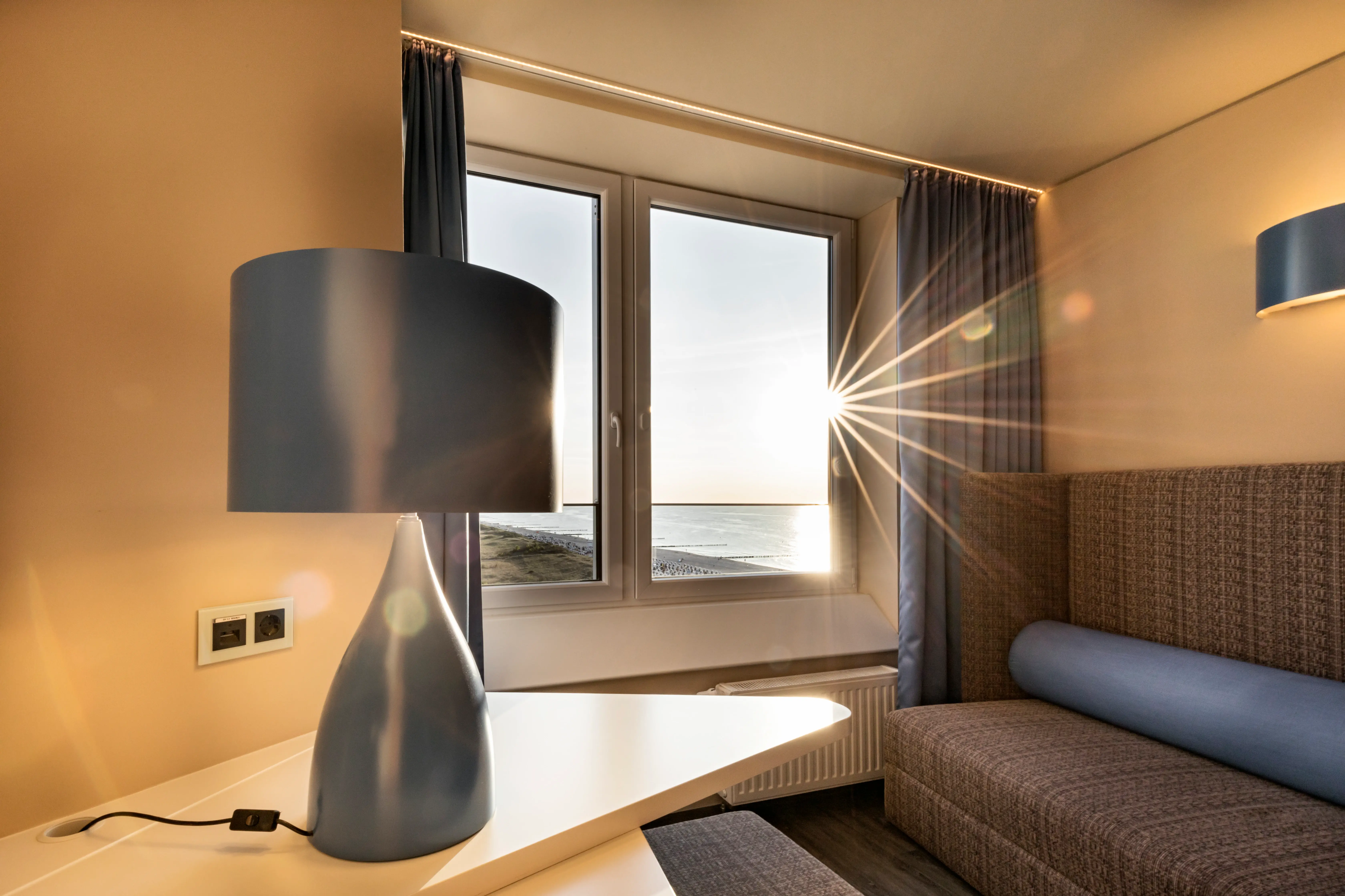 Cozy hotel room with a modern blue lamp on a desk, a comfortable seating area, and a large window framing a stunning sunset over the sea.