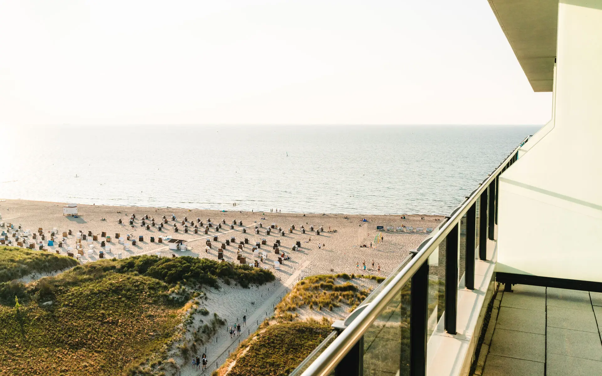 View from a balcony overlooking sandy dunes and a beach with rows of beach chairs, extending to the calm sea under soft sunlight.