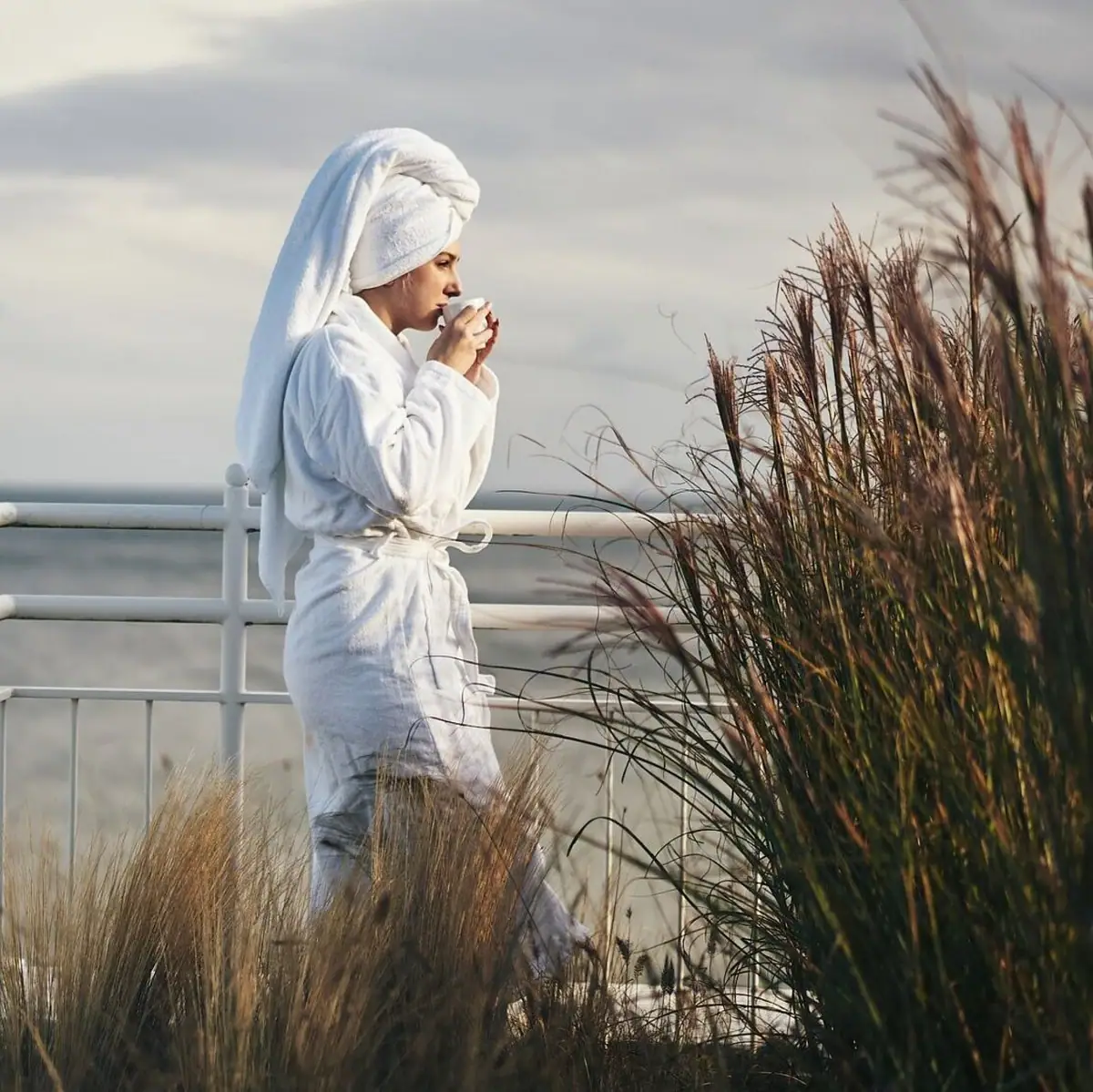 A woman in a bathrobe drinks from a cup.