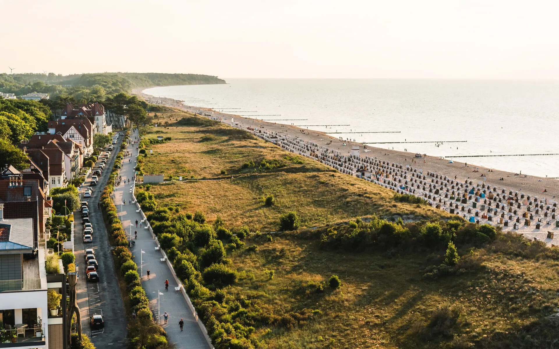 Panoramic view of Warnemünde's beach promenade with charming coastal houses, green dunes, and rows of beach chairs stretching along the Baltic Sea under warm evening sunlight.