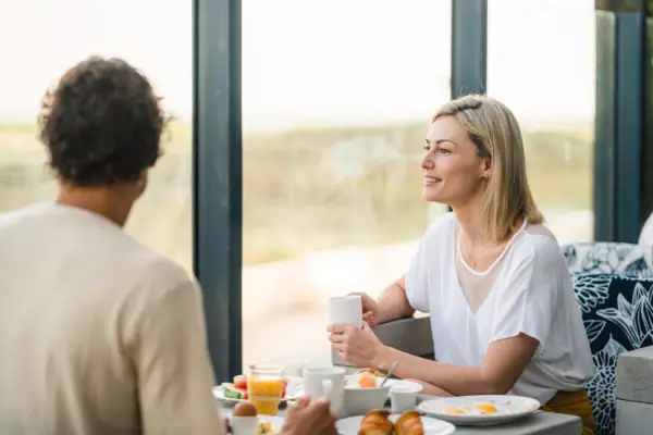 A couple is sitting at the breakfast table and one woman is holding a cup and looking at the other person. The dunes can be seen in the background.