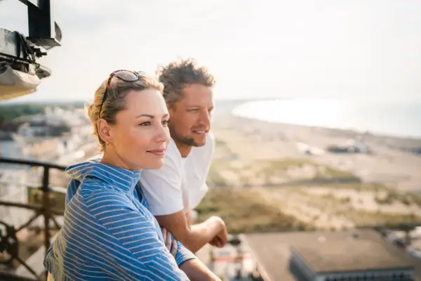 A man and a woman lean against each other on top of the lighthouse railing, smiling and gazing into the distance.