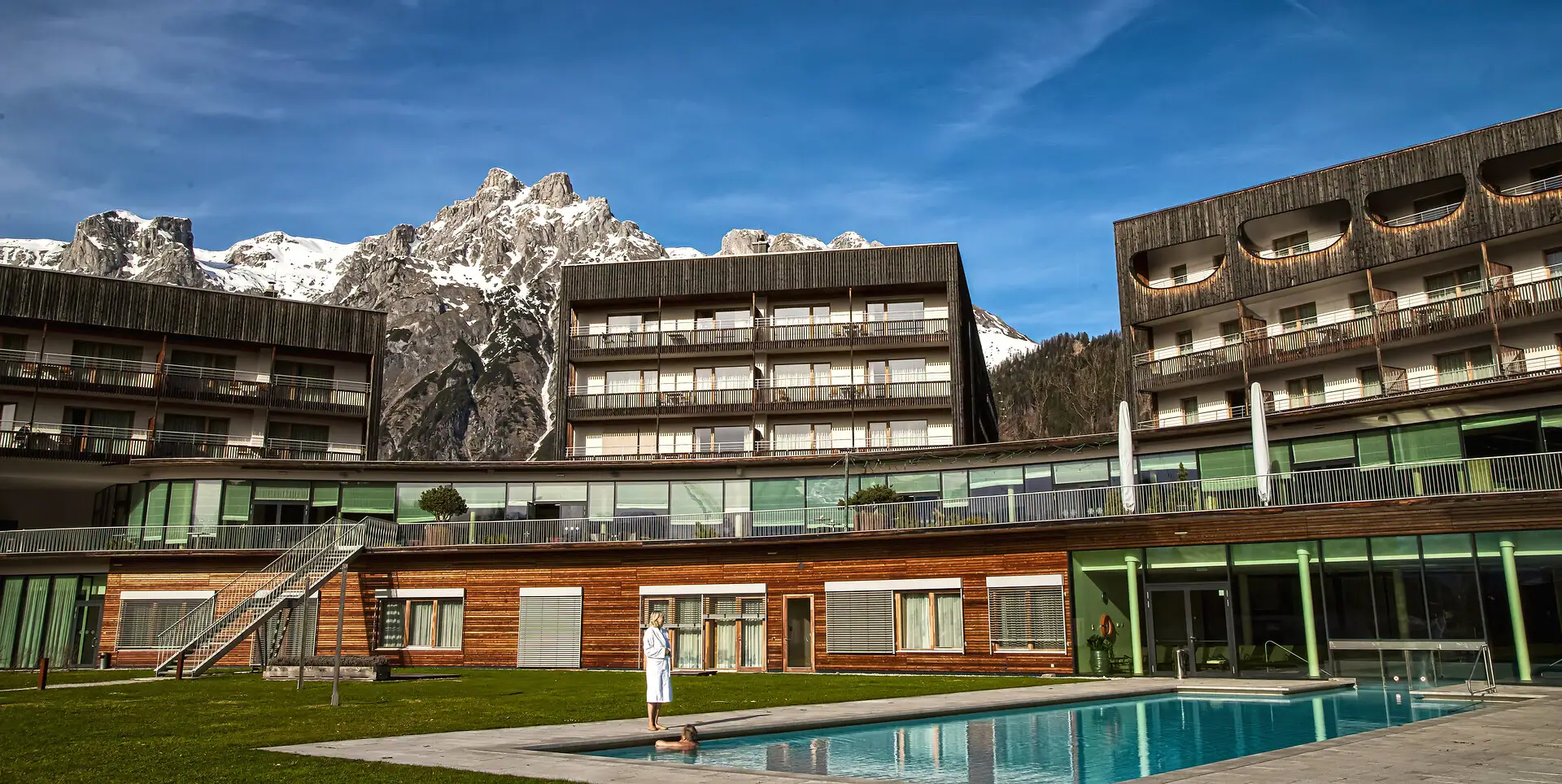 A person is standing in front of a swimming pool and the hotel, outdoors.