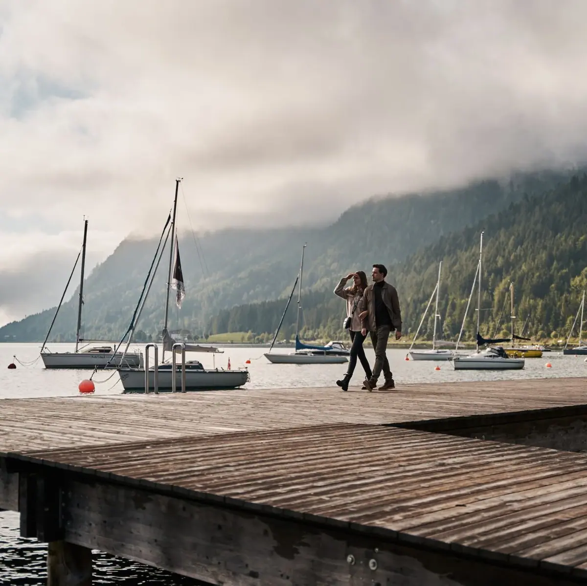 The shores of Lake Achen A couple are walking along a jetty at Lake Achensee, with boats in the background.