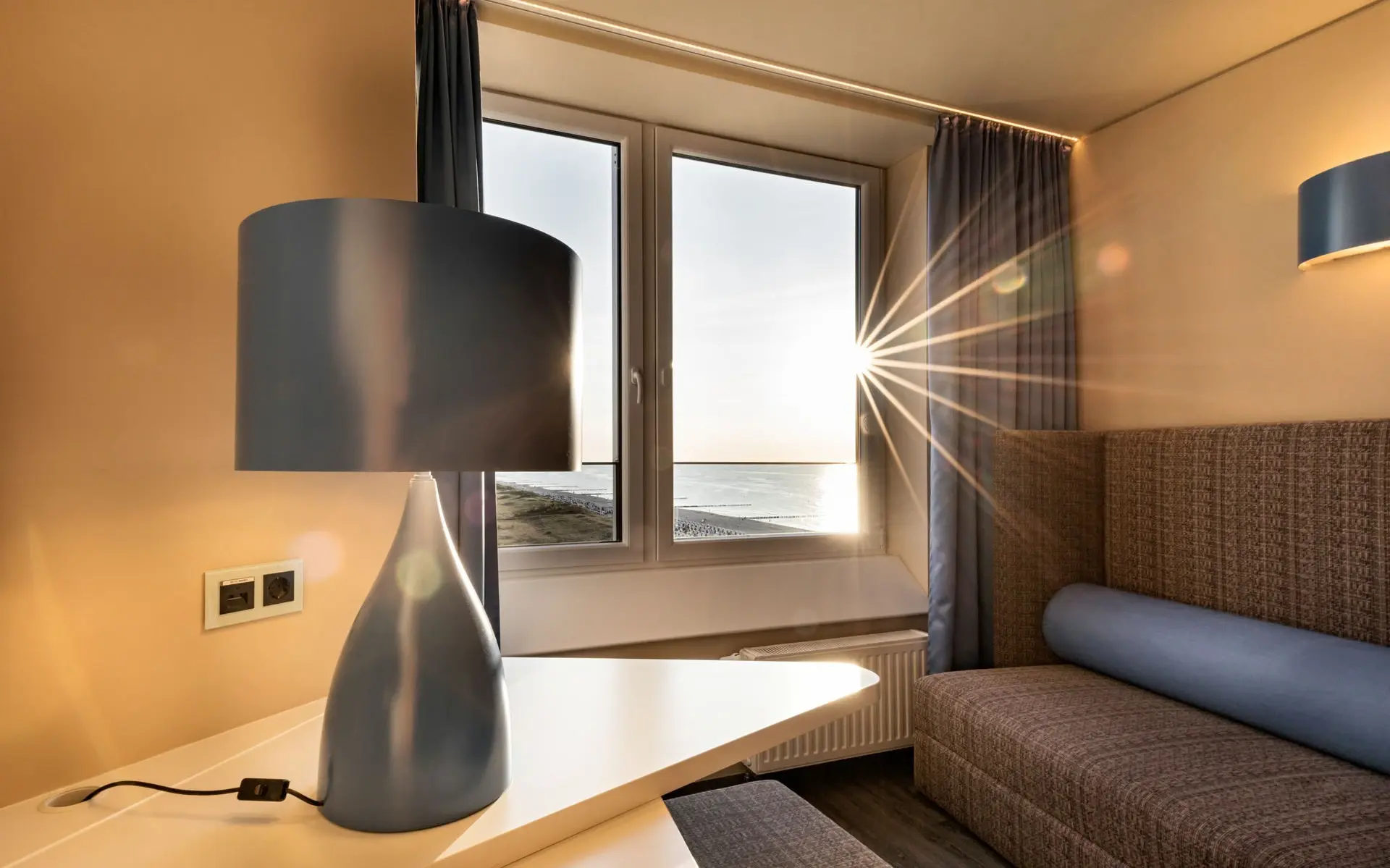 Cozy hotel room with a modern blue lamp on a desk, a comfortable seating area, and a large window framing a stunning sunset over the sea.