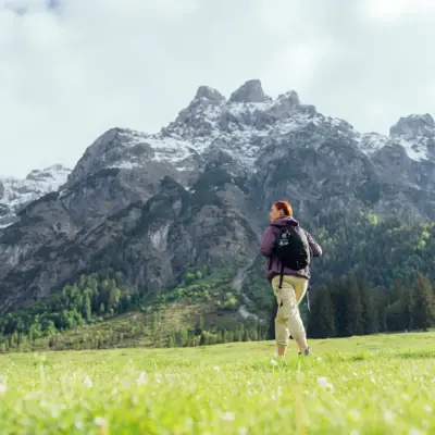A woman standing in a grassy field with mountains and a cloudy sky in the background.