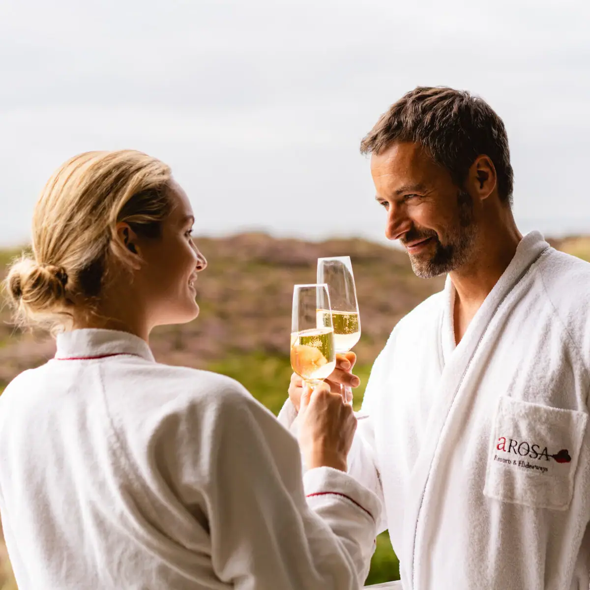 A man and a woman in bathrobes hold wine glasses.