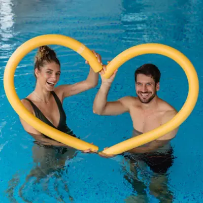 A man and woman in swimwear smiling while holding a yellow tube shaped like a heart in a swimming pool.