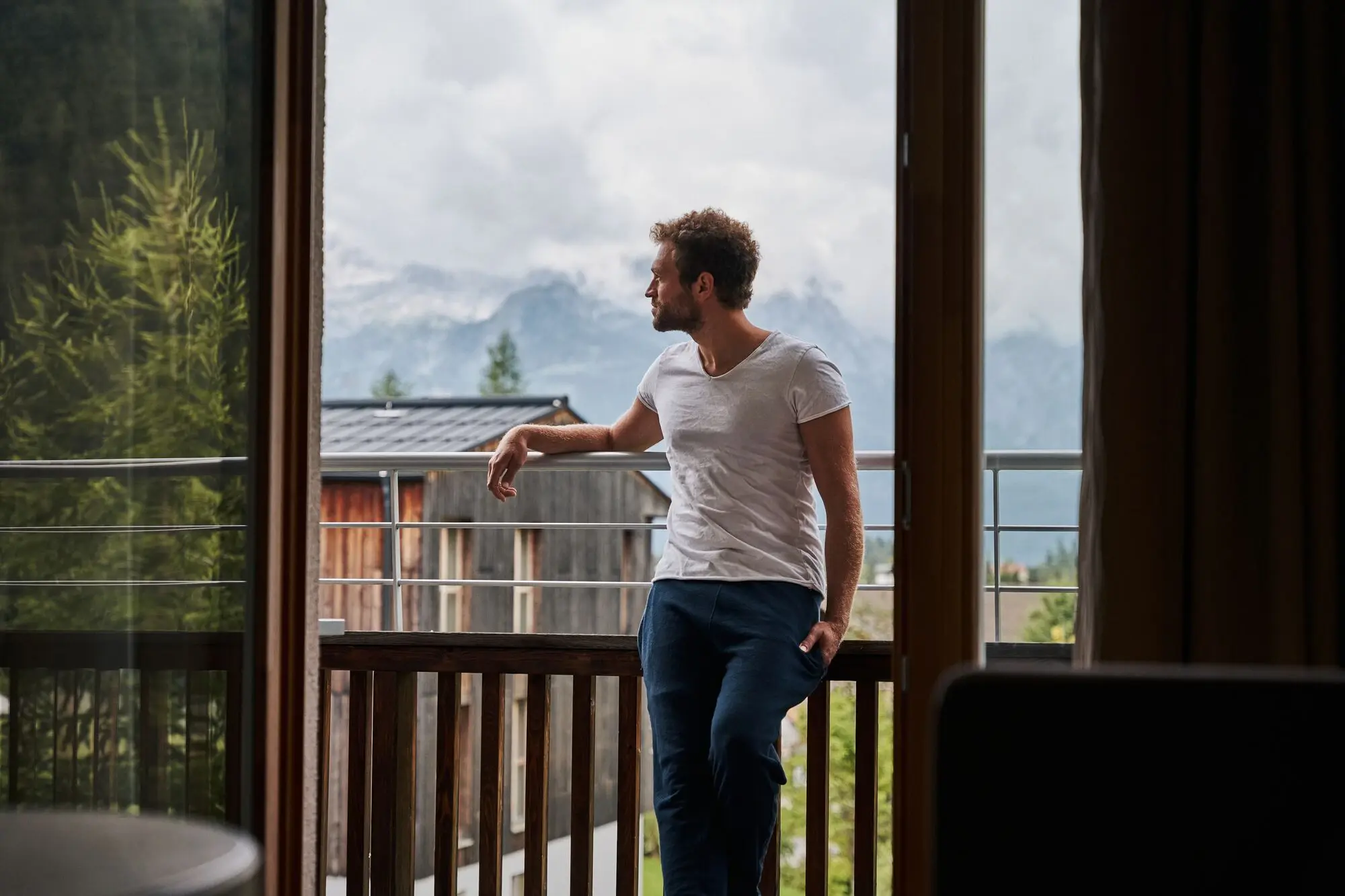 A man leans against a railing and looks out from a balcony onto a mountain range.