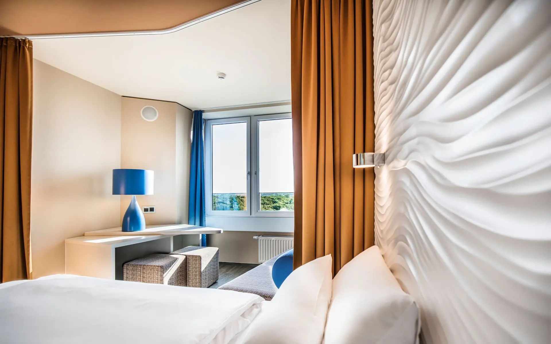 Elegant hotel room featuring a cozy bed with a wave-patterned wall design, a desk with a blue lamp, stylish seating, and a window offering a view of greenery, framed by warm mustard-colored curtains.