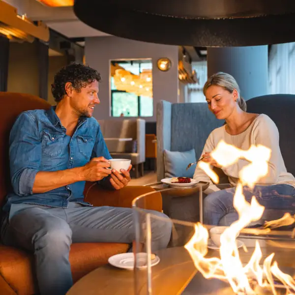 A man and a woman sit on a chair and eat by a fireplace.