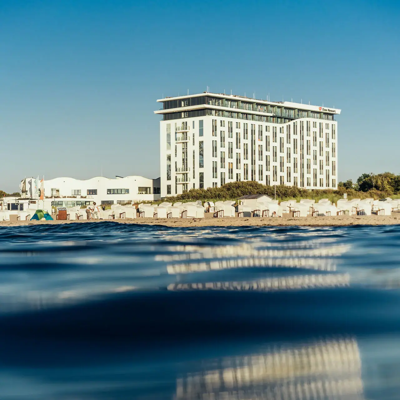 The hotel building on the beach with a clear sky and water in the foreground.
