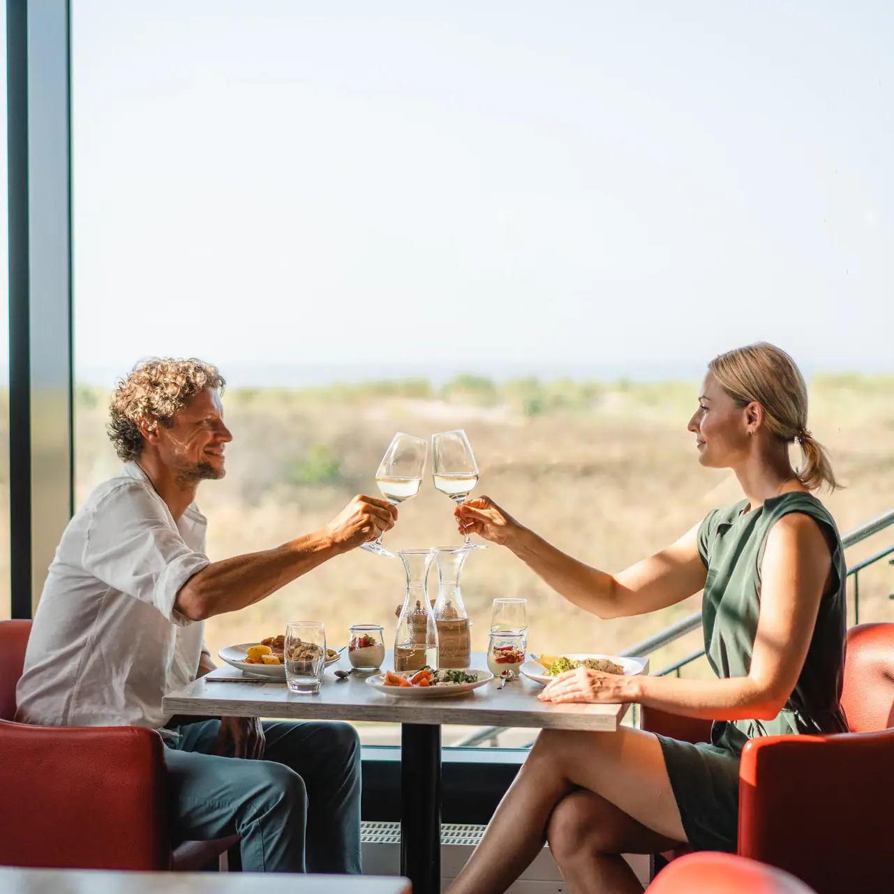 Restaurant at aja Warnemünde A man and a woman are sitting at a table with glasses of wine, with the Baltic Sea in the background.