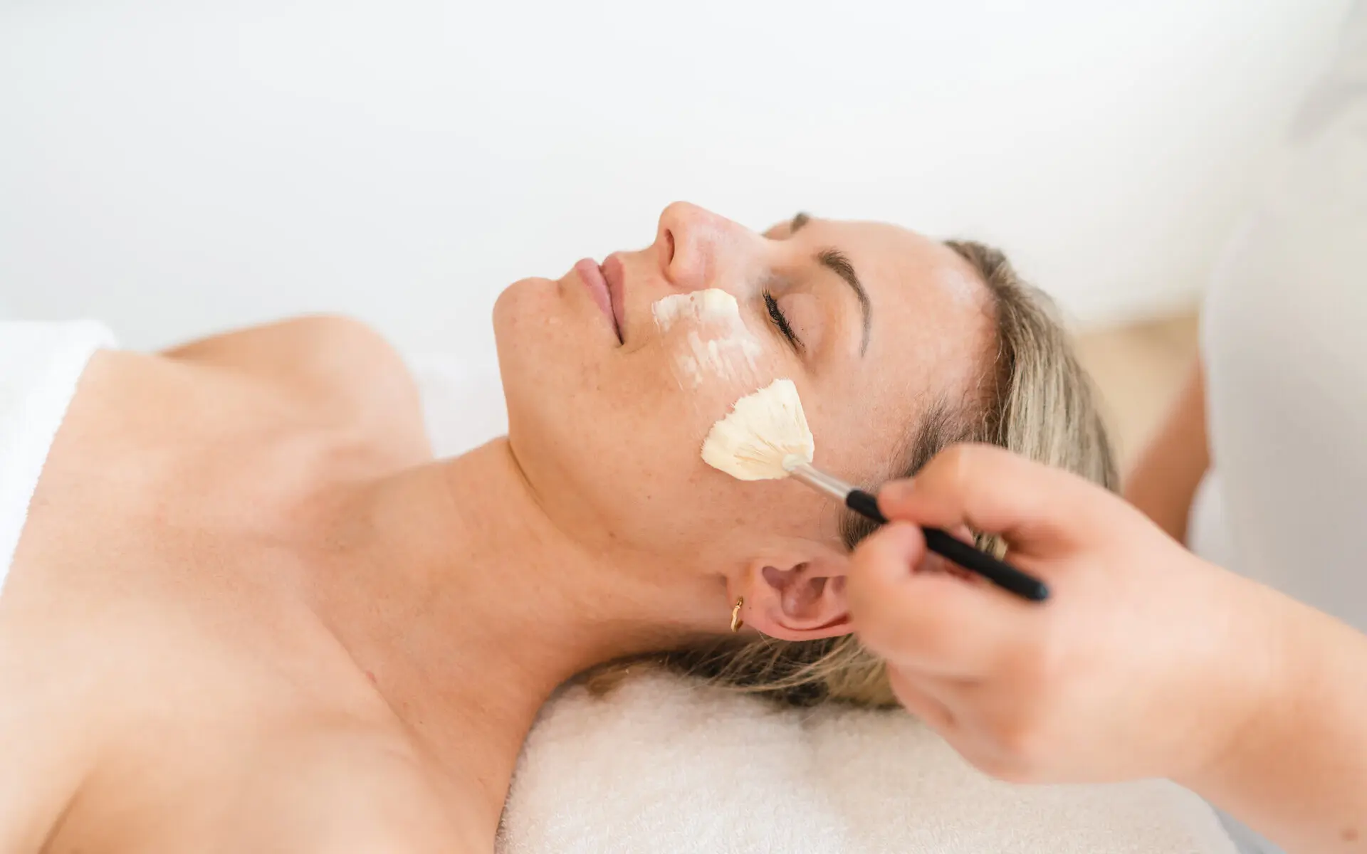 A woman lies relaxed on a lounger and has a face mask applied with a brush.