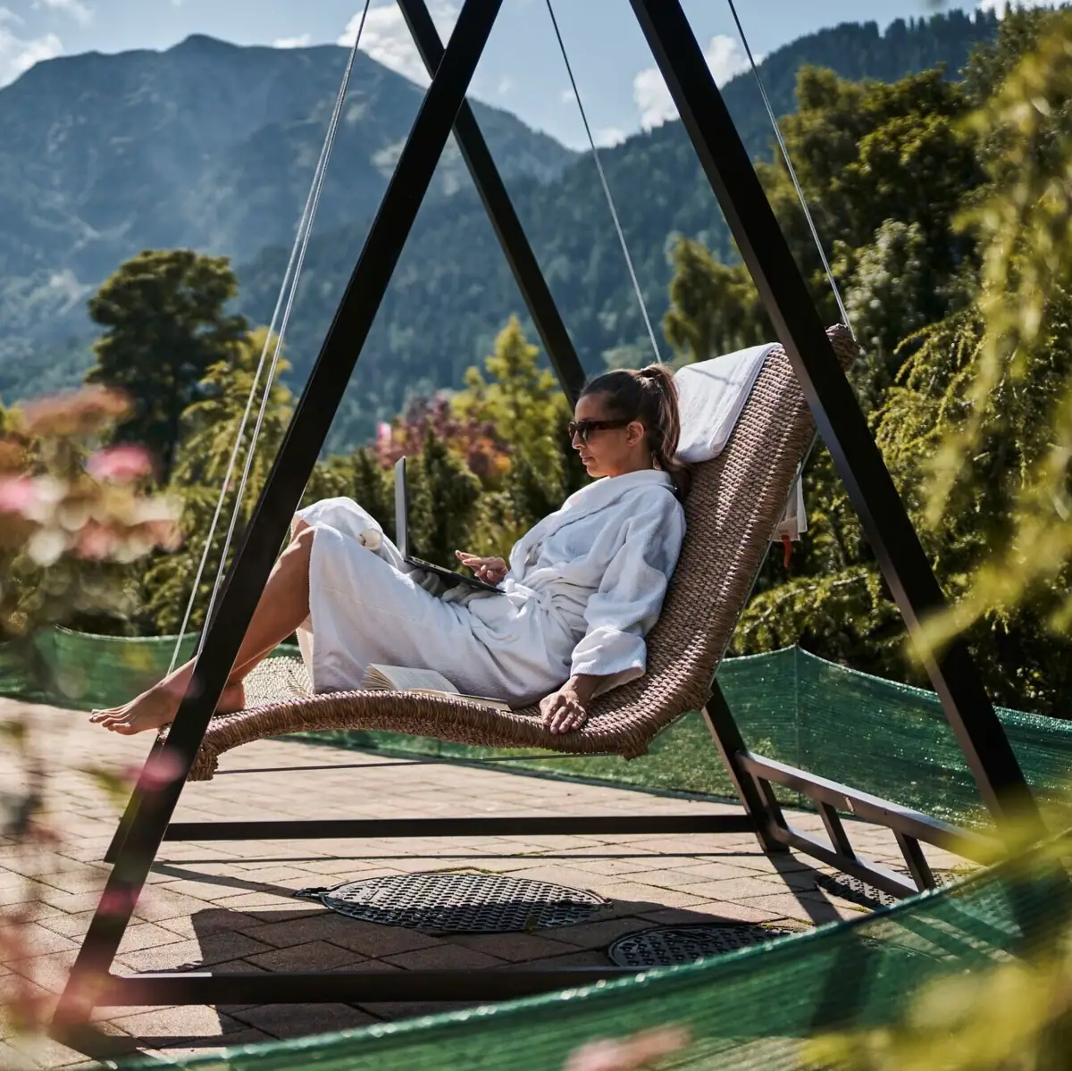 A woman in a white bathrobe sits in a hammock with a laptop.
