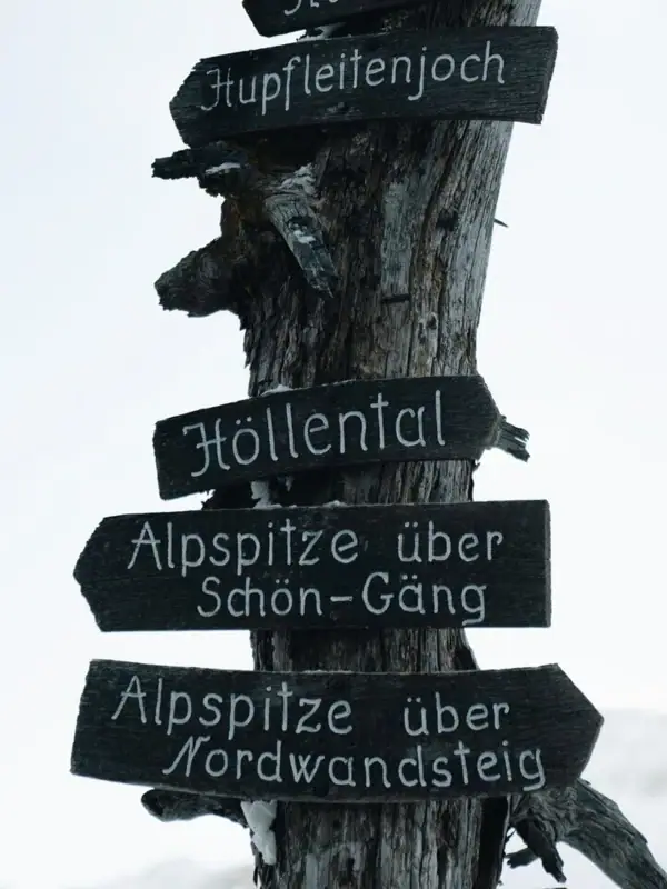 Signpost with many signs in front of a wintry mountain backdrop.