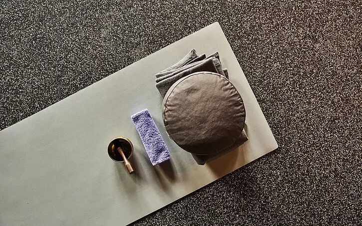 Folded towels, a singing bowl and a round yoga cushion lie on a yoga mat.