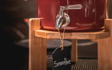 A red smoothie in a drink dispenser on a wooden drink dispenser on a buffet.