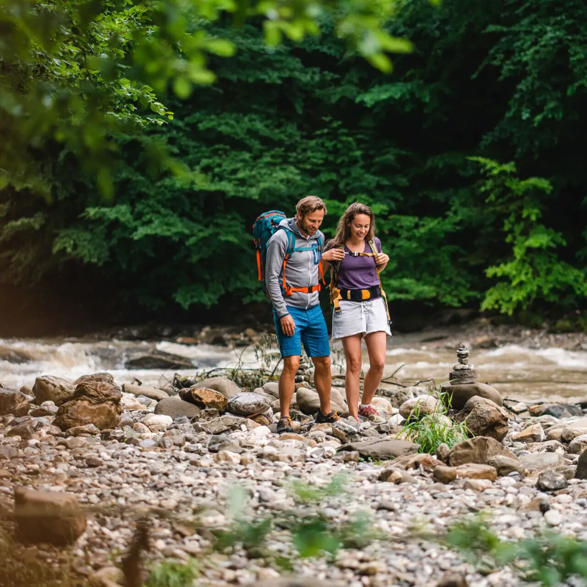 A man and a woman are standing on rocks by a river.