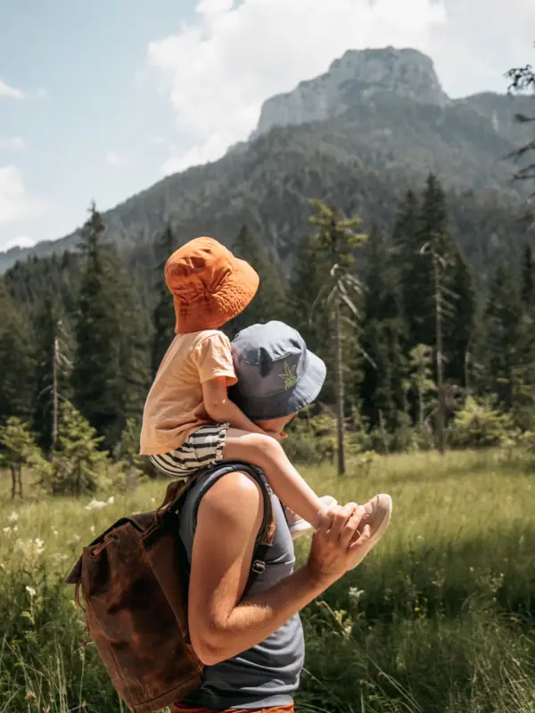 A man is carrying a child on his shoulders in a field with trees and mountains.