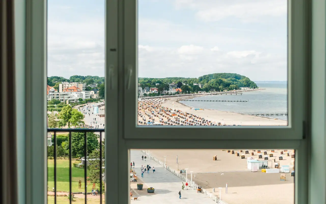 A view through a large window showcasing a sandy beach, beach chairs, and a promenade filled with visitors on a sunny day, framed by the window and a round wooden table in the foreground.