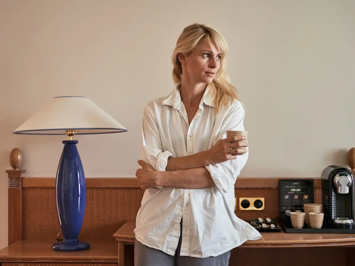 A woman stands in front of a table with a lamp and a coffee cup.