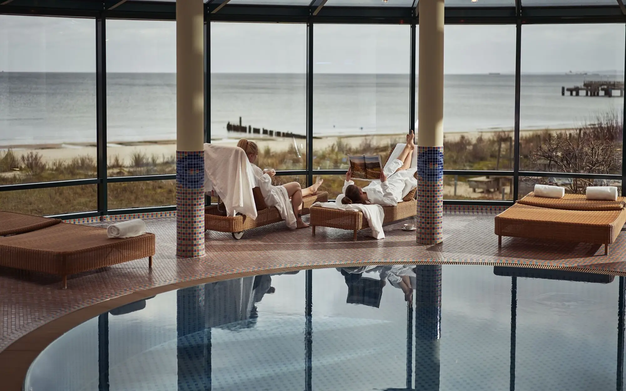 Two people in bathrobes sit by an indoor pool with a sea view.