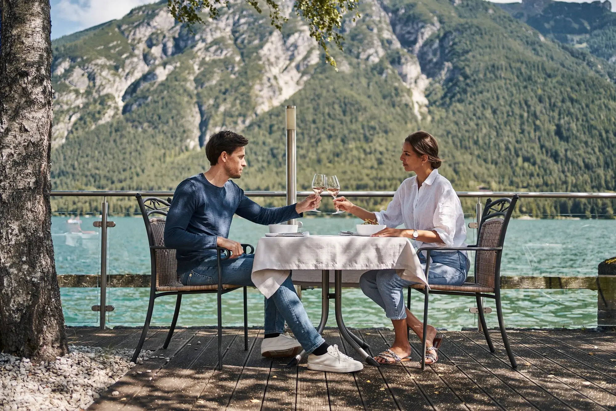 A man and a woman sit at a table and clink glasses of wine, right on the shores of Lake Achensee.