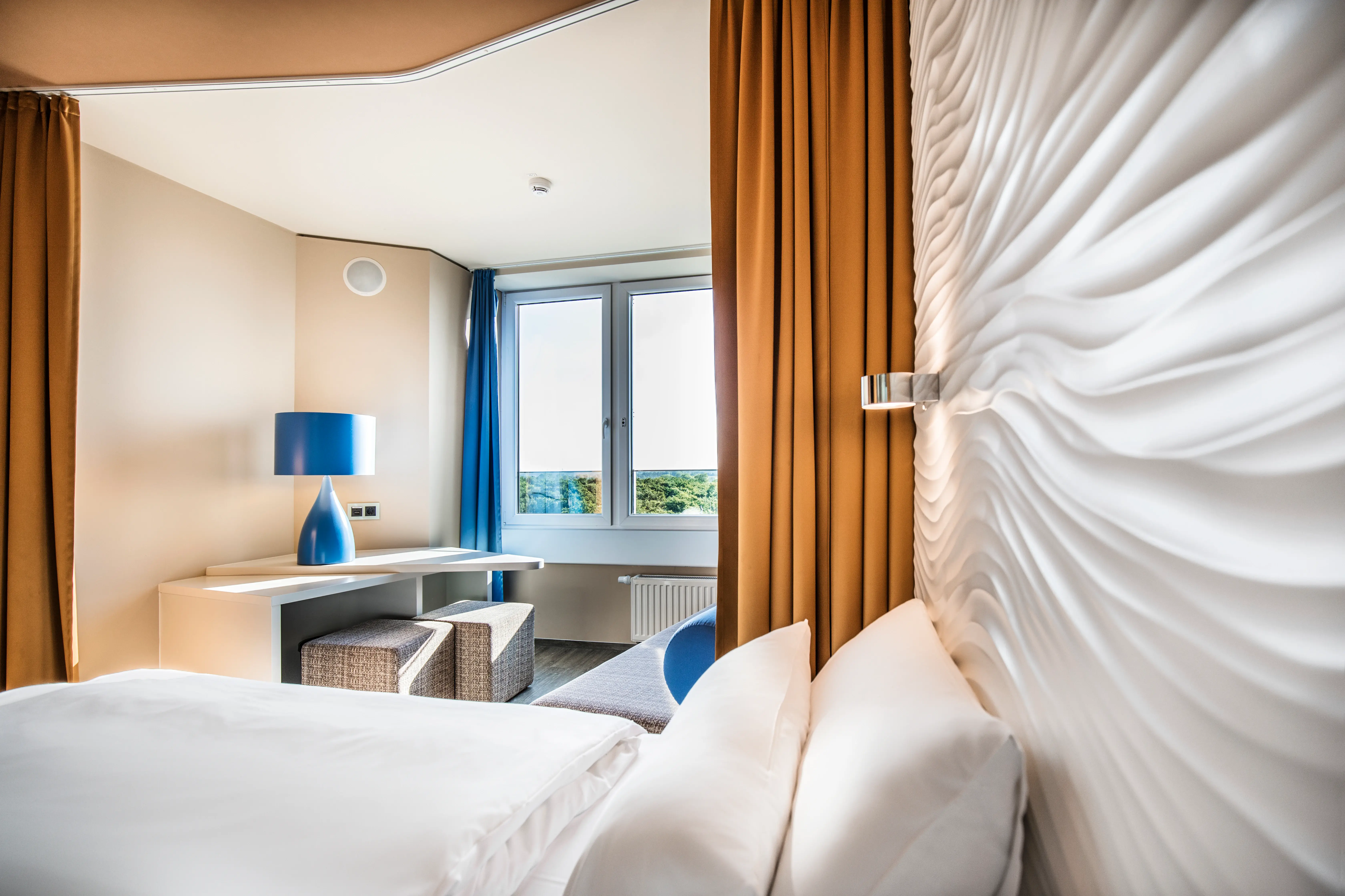 Elegant hotel room featuring a cozy bed with a wave-patterned wall design, a desk with a blue lamp, stylish seating, and a window offering a view of greenery, framed by warm mustard-colored curtains.