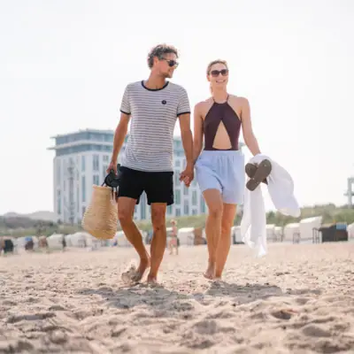 A man and a woman holding hands on the beach, with the aja Warnemünde in the background.