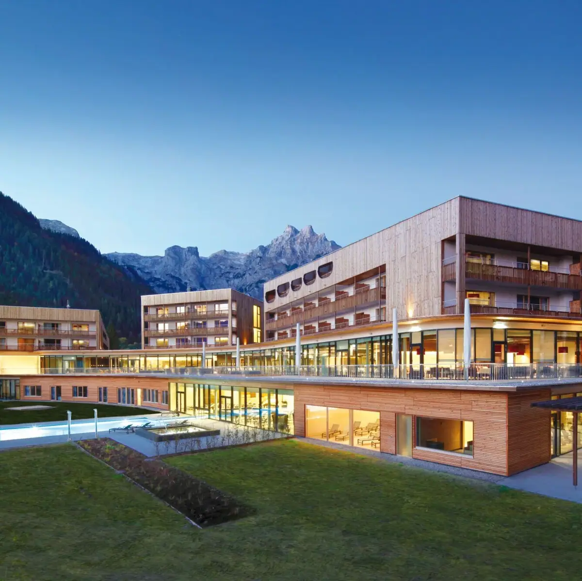 Modern hotel and resort building in Werfenweng, surrounded by an impressive mountain landscape. The architecture combines natural wooden elements with large glass fronts. In the foreground is an illuminated outdoor pool and a spacious terrace with seating.