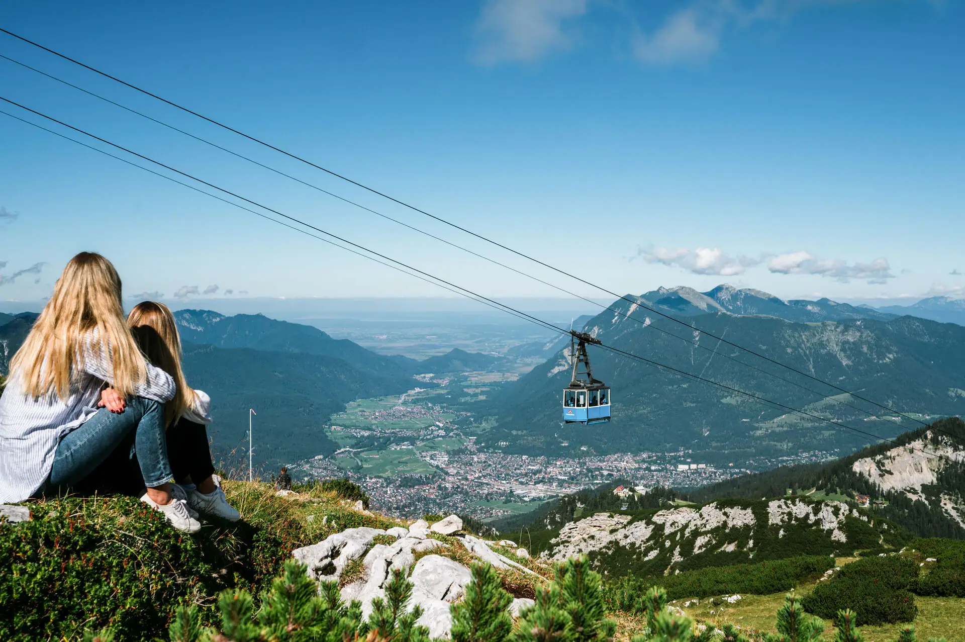 A girl sits on a mountain and looks down into the valley, a cable car hovers above her.